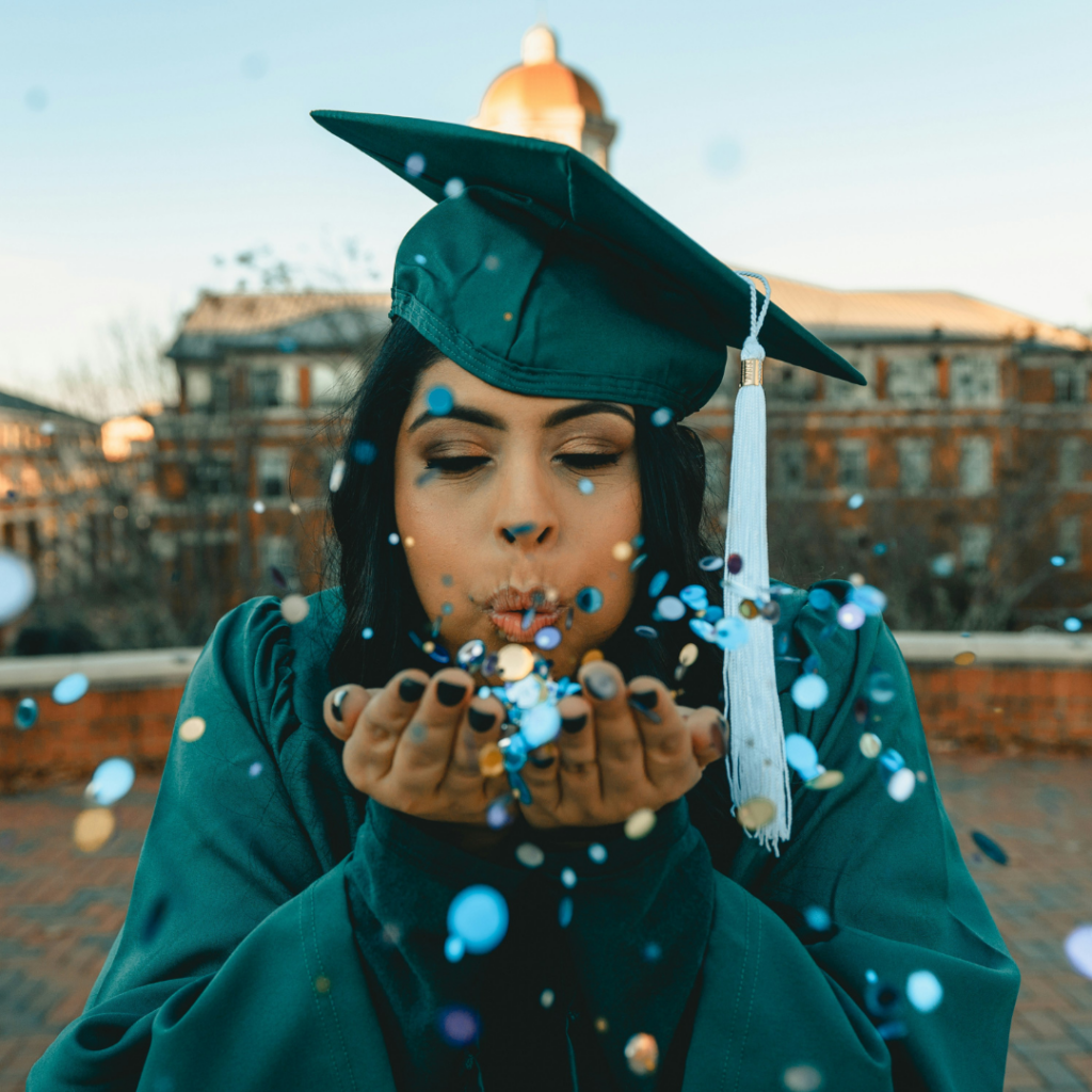 Female graduate blowing confetti to the camera infront of a university building.