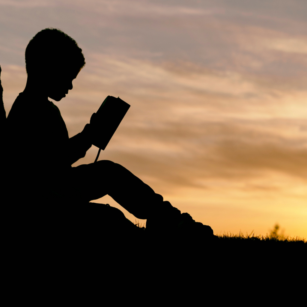 Silhouette of a boy leaned against a tree in a field, reading a book.