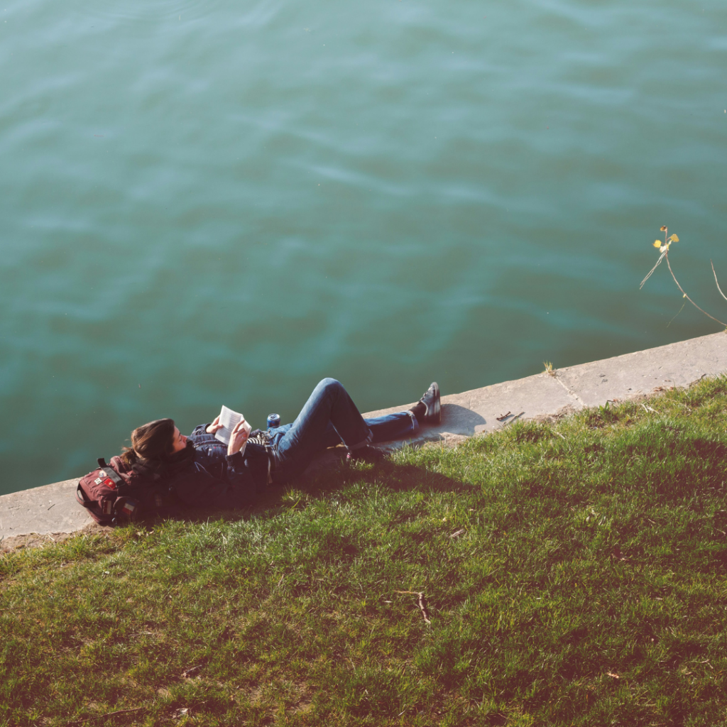 A person laying on the shore next to a river. Reading something, being alone.